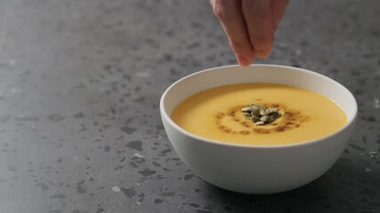 Slow motion pumpkin seeds add to pumpkin cream soup in white bowl on terrazzo countertop - Powered by Adobe