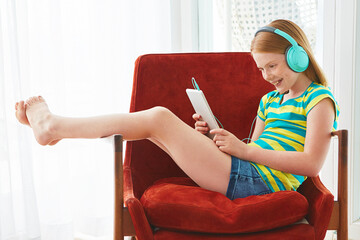 Happy girl, headphones or playing with tablet on chair for online entertainment or music in home. Gamer, teenager or kid with technology, sound app or podcast for streaming or watching in house