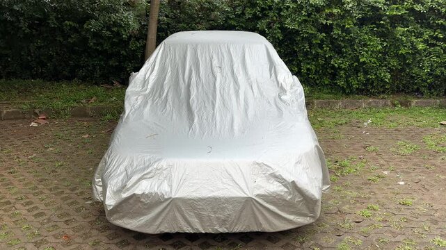Car covered with protective silver tarp in outdoor parking area, vehicle protection from rain, dust, and sunlight, parked automobile fully wrapped with car cover