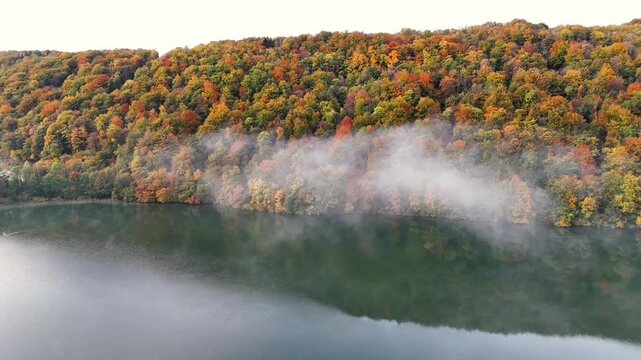 A tranquil autumn scene at Lac du Val in Le Herisson, France, where mist drifts above a calm lake beside a hillside covered in vibrant fall foliage of red, yellow and green trees under natural light