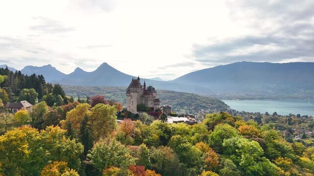 Burg Menthon-Saint-Bernard in France stands above vibrant autumn trees, framed by mountains and a calm lake. The medieval castle dominates the hillside, offering a view of the surrounding landscape