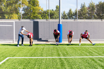 Football players in sports gear warming up and stretching their bodies on a green artificial turf field with their coach
