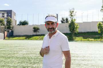 Soccer coach in visor and uniform on sunny outdoor stadium field, confident mature trainer with whistle directing team during training