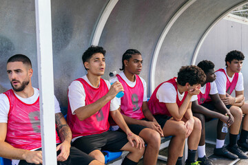 Diverse team of young soccer players watching game from player's bench, some drinking water, waiting to play