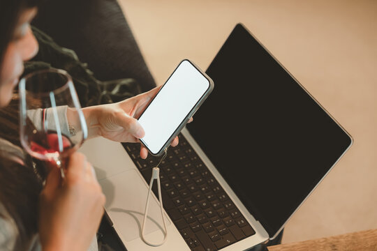 Woman holding smartphone with blank screen and drinking wine beside laptop, representing digital lifestyle,Businesswoman relaxation after work, social media browsing, or modern multitasking at home - Powered by Adobe