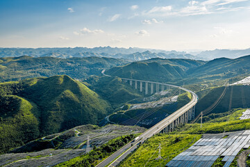 Aerial view of highway viaduct crossing green mountains with solar panels on slopes.
