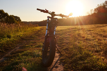 Silhouetted fat tire electric bike parked on a dirt path in a sunny grassy field at sunset, with lens flare emphasizing the golden hour glow and natural landscape backdrop