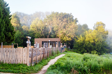 Tranquil morning scene featuring a modern riverside cafe terrace enclosed by a rustic wooden fence leading to lush, misty greenery