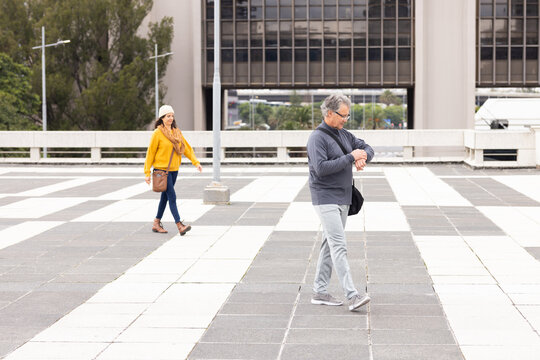 Diverse couple walking plaza past lamp posts and railing checking wristwatch with purse, copy space