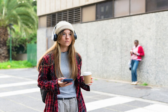 Diverse women walking toward camera and sipping coffee on city sidewalk with smartphone, backpack