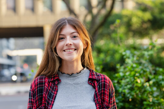 Woman in mid-twenties wearing plaid shirt smiling on sidewalk near building and shrubs