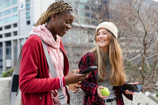 Diverse female friends standing on ledge in plaza holding smartphones, green apple and coffee cup