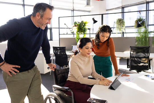 Diverse coworkers collaborating at table in office with wheelchair user using tablet computer