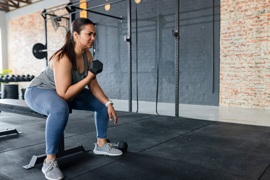 Female athlete performing seated dumbbell bicep curl on bench at gym weight area, copy space