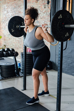 African American woman holding loaded barbell across shoulders in squat rack against brick wall