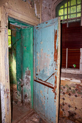 Peeling blue wooden door in a dilapidated room with distressed green walls and arched window light filtering through overgrown trees