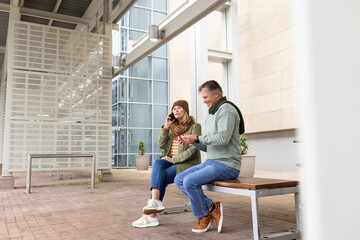 Senior couple sitting on wooden metal-framed bench in outdoor plaza holding smartphones by plants