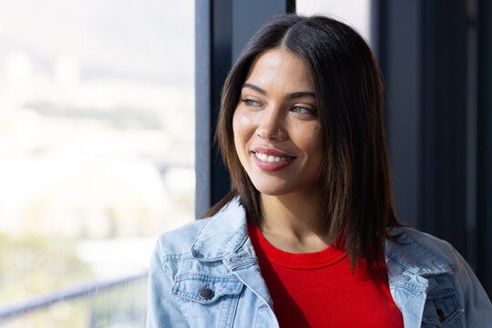 Indian woman gazing at blurred cityscape by window wearing denim jacket, red shirt, copy space
