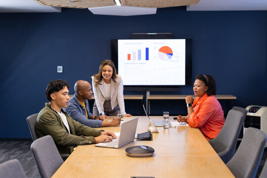 Diverse coworkers sitting around table in conference room using laptops and viewing chart display - Powered by Adobe
