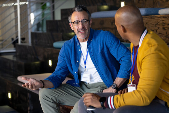 Diverse male colleagues discussing in office lounge, holding coffee cup and wearing lanyard badge