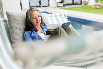 Senior woman in denim shirt reclining in striped hammock beside pool on lawn reading paperback book