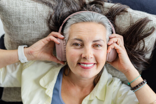 Senior woman reclining on sofa at home fitting pastel-pink headphones with smartwatch and bracelets