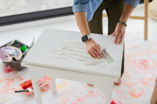 Woman spreading white compound with metal putty knife across wooden table in bright workshop