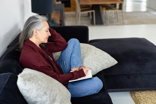 Senior woman sitting on dark grey sectional sofa writing in notebook in modern living room