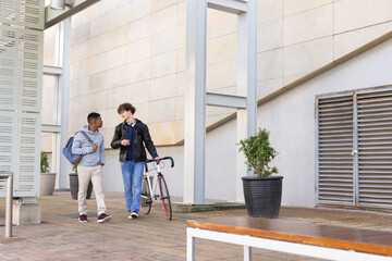 Diverse male friends strolling campus path pushing road bicycle carrying backpack and headphones