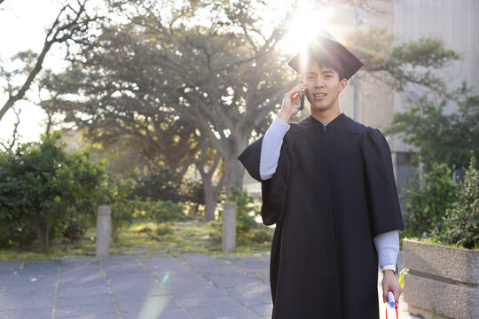 Talking on phone, Asian man on campus path wearing gown cap holding rolled diploma, copy space
