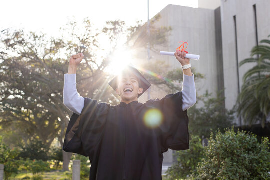 Graduate raising mortarboard and diploma scroll on landscaped campus lawn, with sunlight flaring