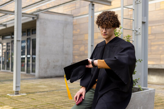 Male graduate wearing gown sitting on courtyard planter holding cap checking smartwatch, copy space
