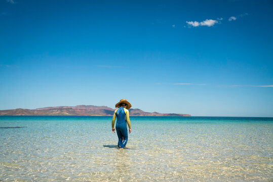 A young girl in wetsuit and sun hat walks in a shallow, clear sea.