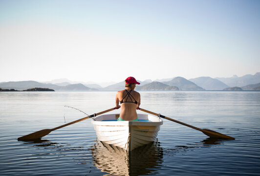 A woman rows a white boat in calm water with mountains in background