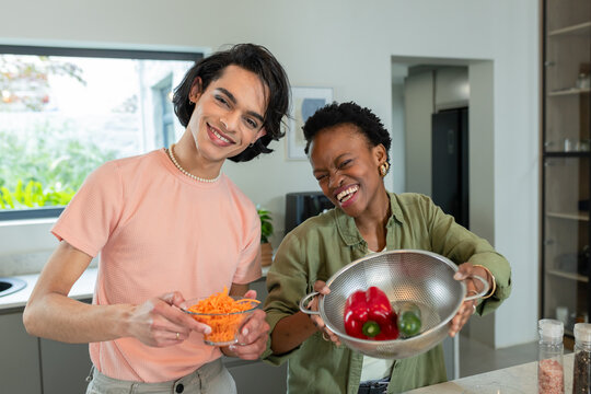 Diverse friends smiling while holding carrots bowl and pepper colander in kitchen on countertop