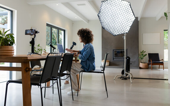 Woman sitting at wooden table creating content with laptop and smartphone under softbox light
