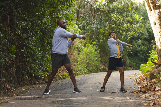 African American father and son stretching arms on paved forest path in athletic shoes - Powered by Adobe