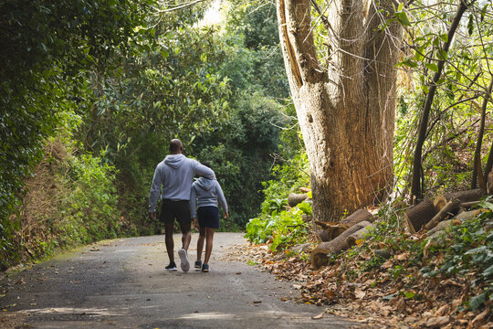 African American father and son walking on paved trail in woods past stacked logs, shrubs, leaves