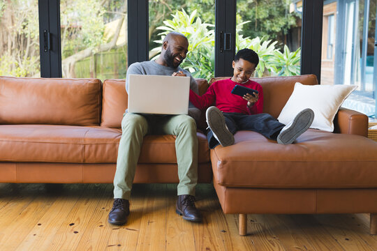 African American father and son browsing laptop and smartphone on sofa beneath living room windows - Powered by Adobe