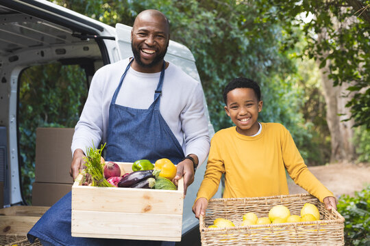 African American father and son holding vegetable crate plus lemon basket beside cargo van at park