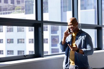 African American man talking on smartphone standing by office windows wearing sweater, copy space