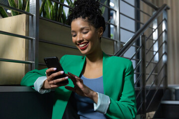 African American woman sitting on office lounge stairs using smartphone beside planter box greenery