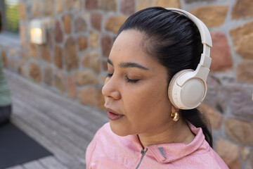 Hispanic woman standing on patio next to stone wall wearing headphones, pink jacket, gold hoops