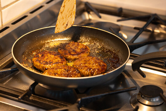 Four seared meat patties are sizzling in non-stick pan on gas stovetop, spatula hovering