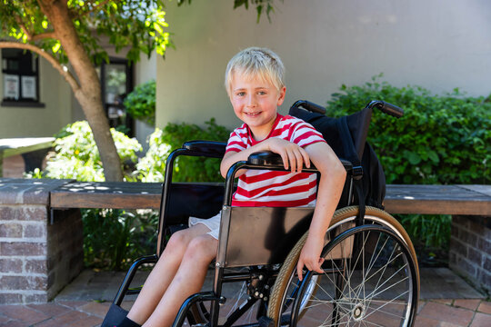 Boy child sitting in wheelchair touching wheel by brick bench in courtyard wearing striped shirt