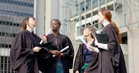After lifting diplomas and passing cap, Women graduates in gowns laughing on plaza, copy space