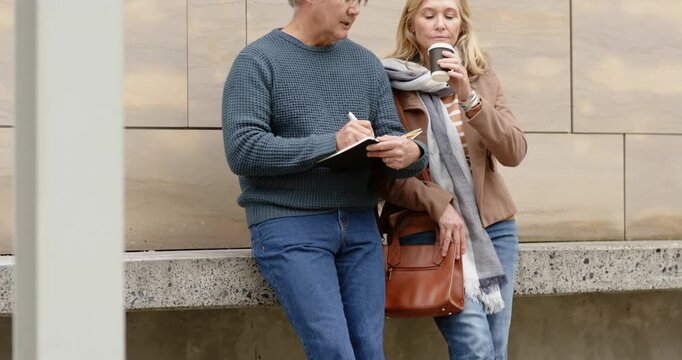 Senior couple strolling to ledge by beige building leaning scribbling notes and sipping coffee
