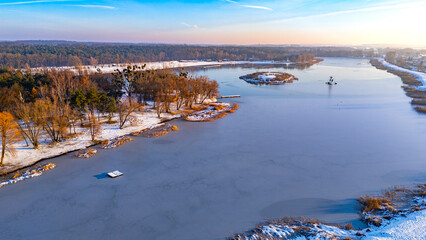 Aerial view of a winter lake with snowy shore, frozen surface, vegetation, small island, pier and surrounding forest landscape