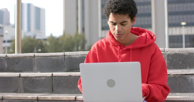 After placing laptop, African American man in red hoodie typing reviewing content on urban steps