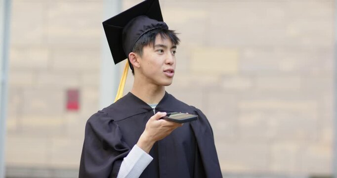 Asian man holding device and wearing cap and gown at campus practicing graduation speech